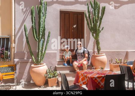 Familie, die auf Stufen vor einem charmanten Haus mit großen Topfkakteen sitzt. Warmes Sonnenlicht erhellt die Terrasse und verbessert die fröhliche Atmosphäre Stockfoto