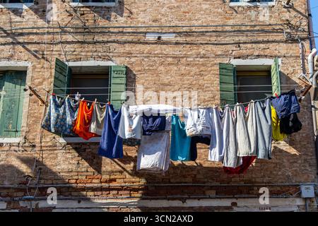 Wäschetrocknung auf Wäscheleinen in der Gasse von Venedig. Bunte Kleidung und Handtücher hängen an einer Schlange zwischen alten Backsteinhäusern mit grünen Fensterläden in einer sonnigen Sonne Stockfoto