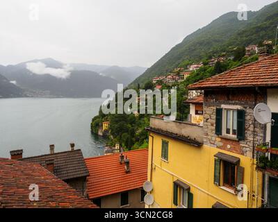 Eine ruhige Aussicht auf den Comer See, umgeben von Hügeln und üppigem Grün, mit lebhaften Ziegeldächern im Vordergrund, die die Region erfassen Stockfoto