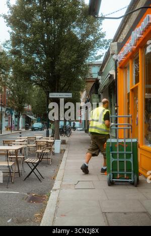 Chiswick, London, Großbritannien – 2025.08.22 – Ein Mitarbeiter in einer gut sichtbaren Weste schiebt einen Wagen an Geschäften und Cafés im Freien auf dem Prince of Wales T vorbei Stockfoto