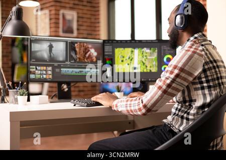 Der afroamerikanische Sounddesigner trägt Kopfhörer, arbeitet von zu Hause aus mit zwei Monitoren, stellt Rohmaterial zusammen und trifft kreative Postproduktionsentscheidungen. Bearbeitung des Workflows, der auf Computerbildschirmen angezeigt wird Stockfoto
