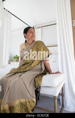 Indische Frau mit beigem Saree, die auf einer weißen Bank neben dem Himmelbett und der Topfpflanze sitzt Stockfoto