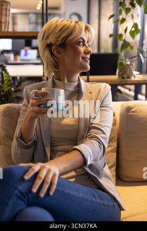 Frau, die auf der Couch in der modernen Bürolounge sitzt und einen zweifarbigen Keramikbecher hält und nach rechts blickt Stockfoto