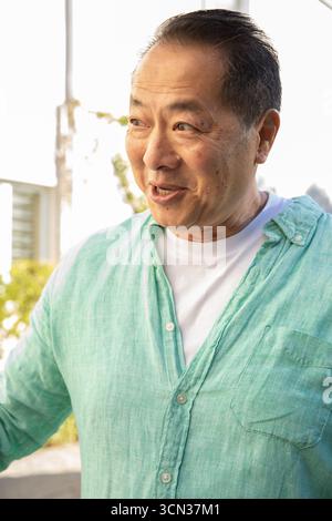 Reifer asiatischer Mann, der auf dem sonnigen Balkon steht und ein grünes Leinenhemd über dem T-Stück trägt und nach links blickt Stockfoto