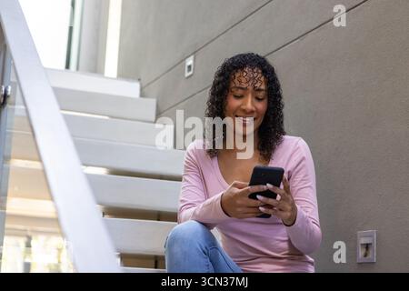 afroamerikanerin sitzt auf einer schwimmenden Treppe am Glasgeländer mit Smartphone, Kopierraum Stockfoto