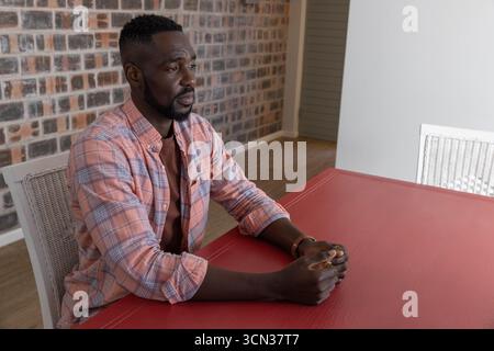 afroamerikanischer Mann sitzt auf weißem Korbstuhl vor der freiliegenden Ziegelwand und blickt auf den Tisch Stockfoto