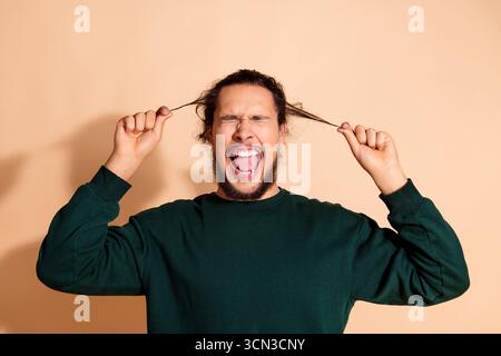 Junger Mann in grünem Pullover, der begeistert Haarsträhnen zieht und Freude vor beigem Hintergrund ausdrückt Stockfoto