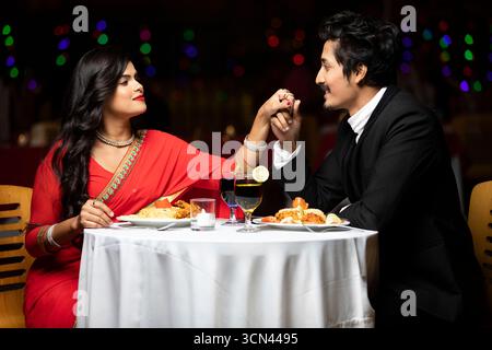 Ein gutaussehendes Paar in traditioneller Kleidung hält Hände und lächelt sich bei einem romantischen Abendessen in einem Restaurant mit warmen Lichtern im an Stockfoto