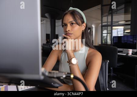 Erwachsene asiatische Frau, die am Schreibtisch im Büro sitzt und auf den Computermonitor mit Kaffeetasse blickt Stockfoto