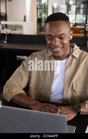 afroamerikanischer Mann, der am Schreibtisch sitzt und einen Laptop benutzt, während er Ohrstöpsel und Armbanduhr trägt Stockfoto