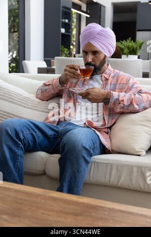 Asiatischer indischer Mann schlürft Tee aus Glas-Teetasse auf Sofa im Wohnzimmer Stockfoto