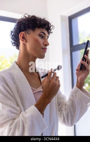 Asiatischer Mann, der Puder mit einem Make-up-Pinsel aufträgt und das Smartphone im Bademantel hält Stockfoto