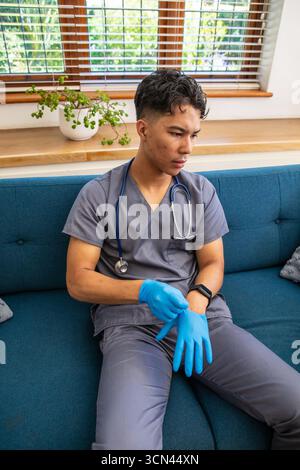 Asiatischer Arbeiter in Peelings mit Stethoskop, der auf Sofa sitzt und Wegwerfhandschuh zieht Stockfoto