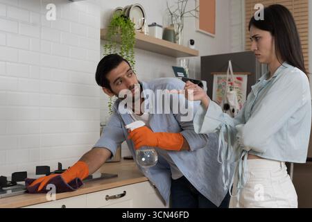 Frustriertes Paar in der Küche, Frau, die mit dem Finger auf den Mann zeigt, der orangefarbene Handschuhe trägt und die Sprühflasche hält, während er den Herd mit einem Tuch reinigt, Konzept von r Stockfoto