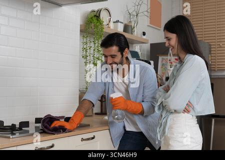 Fröhliches Paar, das die Küche zusammen putzt, Mann mit orangefarbenen Handschuhen, der das Kochfeld mit einem Tuch abwischt, während er die Sprühflasche hält, Frau lächelt und beobachtet Stockfoto