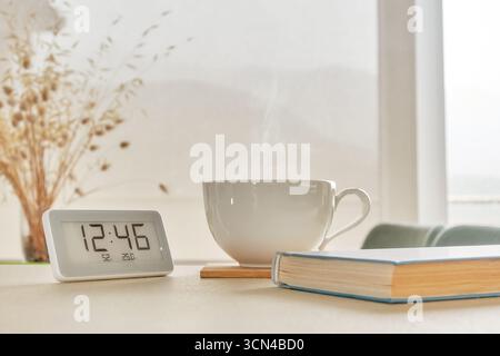 Eine gemütliche Morgenszene mit einer dampfenden Tasse Kaffee neben einem Buch und einer Digitaluhr mit weichem Tageslicht Stockfoto