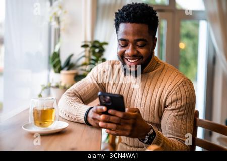 Ein lächelnder schwarzer Mann sitzt an der Bar, lässig gekleidet. Er nutzt Smartphone und Messaging online. Stockfoto