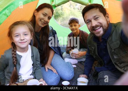 Glückliche Familie, die Selfie im Campingzelt macht, mit Tassen mit heißem Getränk draußen Stockfoto