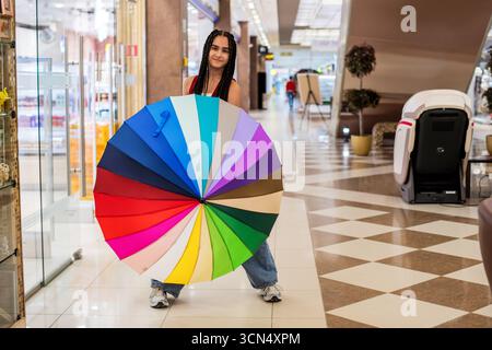 Mädchen mit Zöpfen hält Regenbogenschirm im Einkaufszentrum. Stockfoto