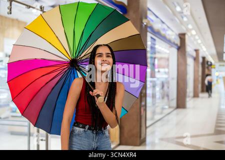 Mädchen mit Zöpfen hält Regenbogenschirm im Einkaufszentrum. Stockfoto