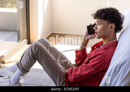 Mann, der im Zimmer sitzt und auf dem Smartphone neben dem Sofa durch eine Glasschiebetür spricht, Kopierraum Stockfoto