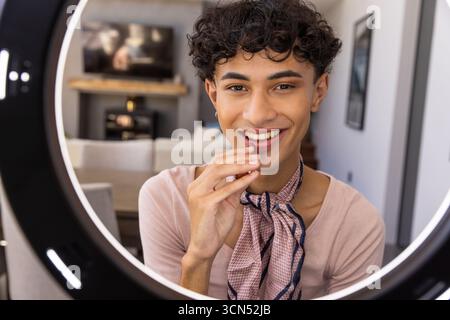 Mann Mitte Zwanziger, der Schal trägt, lächelt in kreisförmiges LED-Ringlicht im Wohnzimmer Stockfoto