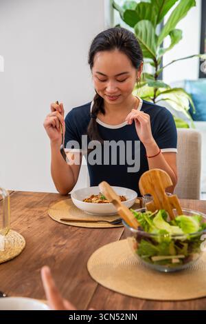 Asiatische chinesin, die mit gewebten Tischsets am Holztisch sitzt und Nudeln aus der Schüssel isst Stockfoto
