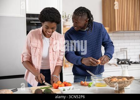 afroamerikanisches Paar, das in der Küche Gemüse hackt und Eier knackt, in der Nähe des Milchbehälters Stockfoto