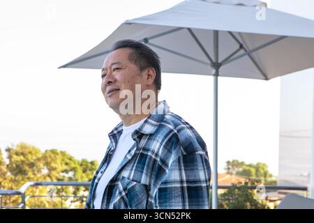 Chinesischer Mann, der auf dem Balkon hinter dem Geländer unter dem Schirm steht und auf die Dächer blickt, Kopierraum Stockfoto