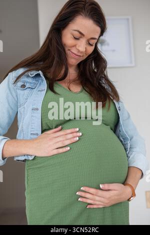 Schwangere Frau, die zu Hause steht und den Bauch in grünem Kleid trägt, Armbanduhr und Halskette Stockfoto