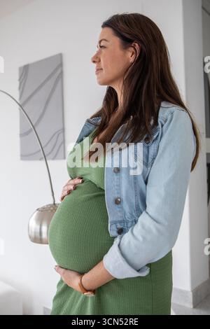 Schwangere Frau, die im Wohnzimmer steht und mit abstrakter Leinwand und Bogenlampe einen Kopierraum betäubt Stockfoto