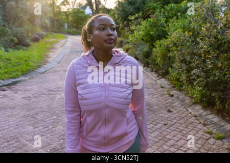 afroamerikanerin, die auf gemauerten Wegen durch den Park geht, trägt Jacke und Ohrstöpsel Stockfoto