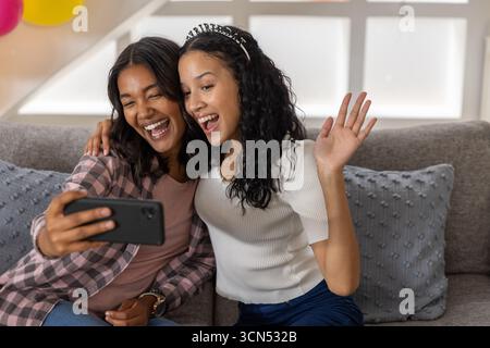 Vielfältige Mutter und Tochter im Teenageralter lächeln, während sie ihr Smartphone halten und zu Hause auf der Couch posieren Stockfoto
