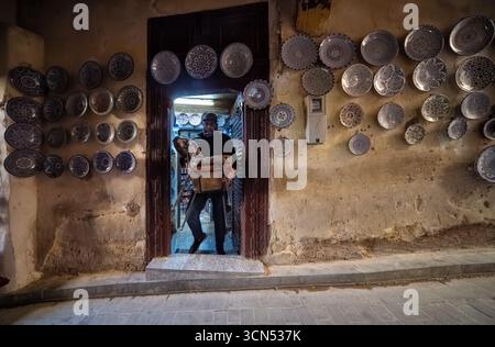 Fès, Marokko, 4. Januar 2023, farbenfrohe Keramikplatten schmücken die Wände eines Ladens in der Medina Fès, während ein Mann Waren vom Eingang trägt. Eine Mischung aus Kult Stockfoto