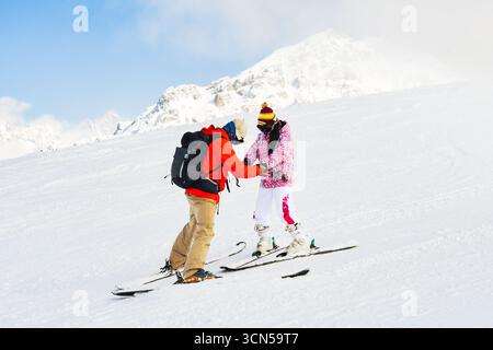 Männlicher Skilehrer in roter Jacke, der die Hände hält, Anfänger weibliche Erwachsene Skifahrerin in rosa Outfit auf verschneiten Berghängen, Konzept der Skischule, Trai Stockfoto