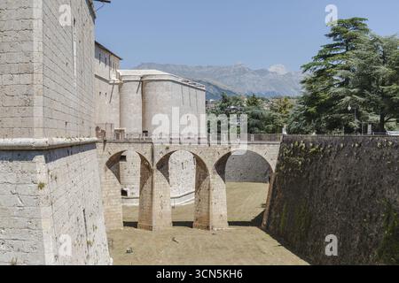 View of ancient stone walls and arched bridge connecting fortress sections under a clear sky, with distant mountains adding depth, L'Aquila, Abruzzo, Stockfoto