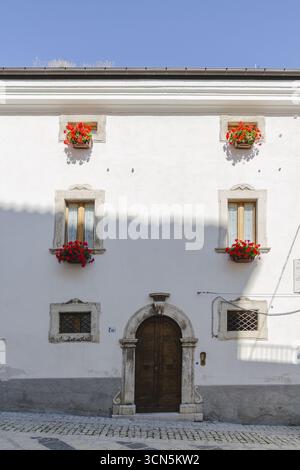 Blick auf eine helle weiße Gebäudefassade mit leuchtenden roten Blumen in Fensterkästen, eingerahmt von einer bogenförmigen Tür, Pescocostanzo, Abruzzen, Italien. Stockfoto