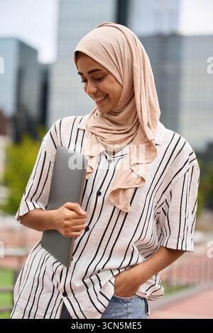 Eine junge muslimische Frau lächelt und hält draußen einen Laptop Stockfoto