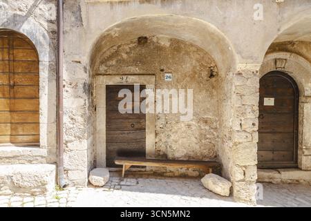 Blick auf alte Steinbögen, die verwitterte Holztüren umrahmen, und eine einfache Bank, die in Sonnenlicht getaucht ist, schaffen eine zeitlose Szene, Santo Stefano di Sessani Stockfoto