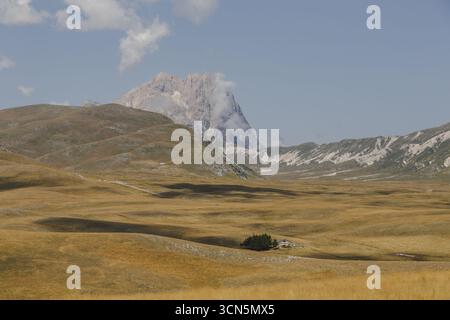 Blick auf goldene Felder erstrecken sich in Richtung des majestätischen Gran Sasso, dessen Gipfel von weichen Wolken bedeckt ist und Schatten in Isola del Gran Sasso D'italia, Abruzzen, Italien wirft. Stockfoto