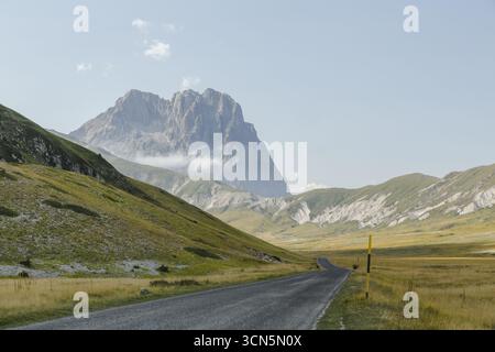 Blick auf die Asphaltstraße, die durch goldene Felder in Richtung der majestätischen Gran Sasso Bergkette unter weichem Himmel führt, Isola del Gran Sasso D'italia, Abruzzen, Italien. Stockfoto