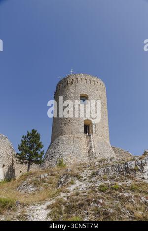Der Blick auf den alten Steinturm erhebt sich majestätisch gegen den klaren blauen Himmel, ein einsamer Baum, der auf dem Hügel wacht, Bominaco, Abruzzen, Italien. Stockfoto