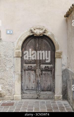 Blick auf eine alte Holztür mit Steinbogen steht verwittert und strukturiert vor einem hellen Gebäude, eingerahmt von Kopfsteinpflaster, Anversa degli Abruzzi, Abruzzen, Italien. Stockfoto