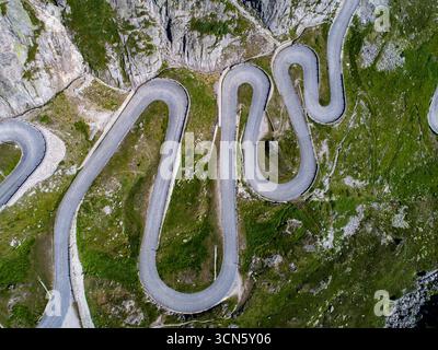 Der Blick auf eine gewundene Straße schlängelt sich durch die zerklüftete, grüne Landschaft und kontrastiert mit den grauen Felswänden, Gotthard Pass, Tessin, Schweiz. Stockfoto
