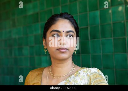 Indische Frau, die an einer grünen Wand steht, in geblümtem Sari mit Goldschmuck und blauem Bindi Stockfoto