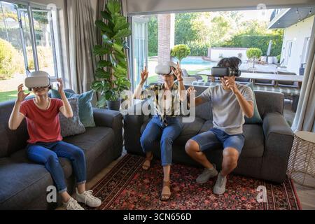 Vielseitige Familie, die in hellem Wohnzimmer auf Sofas sitzt, vr-Headsets trägt und die Hände hebt Stockfoto