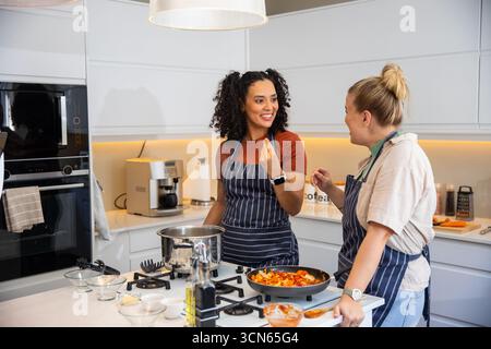 Verschiedene Freundinnen in gestreiften Schürzen kochen, während sie sich auf der Kücheninsel unterhalten Stockfoto