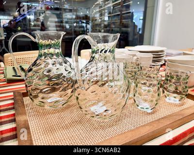Krüge und Becher aus strukturiertem Glas werden auf einem Holztablett wunderschön ausgestellt, wodurch ihr kompliziertes Design hervorgehoben und eine einladende Atmosphäre geschaffen wird Stockfoto