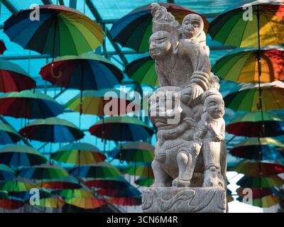 Traditionelle chinesische Statue mit farbenfrohen Regenschirmen - Eine detailgetreue Steinstatue einer traditionellen chinesischen Figur steht unter einer lebendigen Decke aus Farben Stockfoto