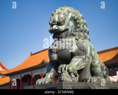 Eine detaillierte, flache Aufnahme einer majestätischen Bronzeblöwen-Statue in der Verbotenen Stadt mit einem kaiserlichen Palastdach im Hintergrund und einem klaren blauen Himmel Stockfoto